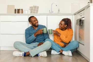 Romantic black couple drinking tea, sitting in kitchen on floor and talking, clinking cups with hot drink and smiling to each other, enjoying morning at home