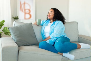 African American Woman Drinking Coffee Holding Mug Sitting On Couch Enjoying Weekend At Home. Lady Resting On Sofa In Modern Living Room. Domestic Comfort Concept