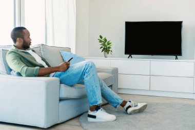 Young black man watching tv with empty screen, resting on sofa in living room interior, mockup. Entertainment with modern technology at spare time, news and new movie