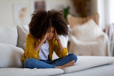 Unhappy little black girl with bushy hair suffering from headache, sitting on couch at home and touching her head, looking at copy space, wearing casual outfit. Kids health care concept