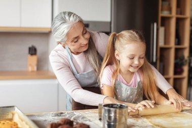 Cheerful caucasian elderly grandmother in apron teaching small girl to make dough with rolling pin for pizza and cookies in kitchen interior. Lesson of prepare homemade food, cooking together at home