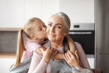 Portrait of smiling european little granddaughter kisses cheek of elderly grandmother in apron in minimalist kitchen interior. Cooking together for family at home, love and relationship, housekeeping