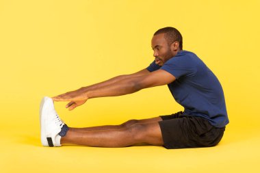 Athletic African American Man Exercising Stretching Touching Toes Sitting Over Yellow Studio Background, Wearing Fitwear. Fitness Workout, Healthy Training Concept. Side View Shot