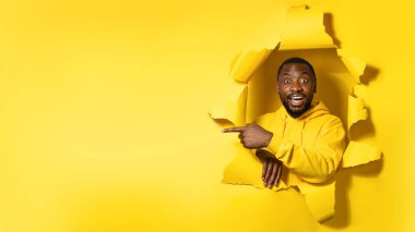 Cheerful black man pointing aside recommending best offer, peeking out through torn paper hole, yellow background, panorama with free space