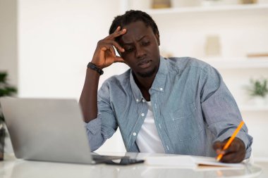 Pensive Black Man Taking Notes While Working With Laptop Computer At Home Office, African American Male Freelancer Sitting At Desk And Writing In Notepad, Noting Down Information, Copy Space