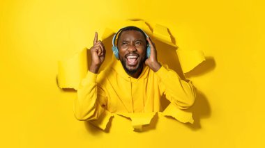 Emotional black man listening music in headphones and singing favourite song, peeking out hole on yellow background, pointing finger up, panorama