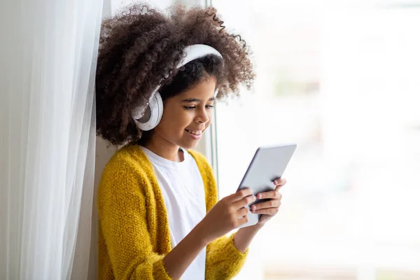 Closeup photo of cheerful black little girl sitting next to window at home, using brand new digital pad and white wireless headset, looking at tablet screen and smiling, watching cartoons, copy space