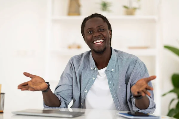 Cheerful young black man sitting at desk and talking at camera, smiling african american male in casual clothes having conversation with somebody, freelancer guy posing at home office, copy space