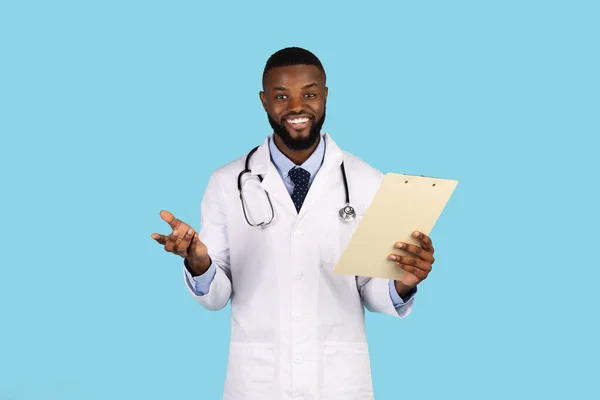 Medical Services. Portrait Of Smiling Black Male Doctor In Uniform With Clipboard In Hand Posing On Blue Background, Friendly African American Physician In White Coat Talking At Camera, Copy Space