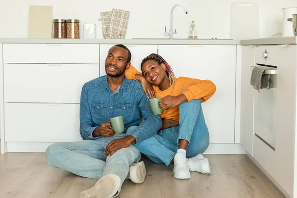 Romantic black couple drinking coffee and looking away, sitting on floor in kitchen, dreaming and talking, enjoying morning at home. Spouses having hot caffeine drinks