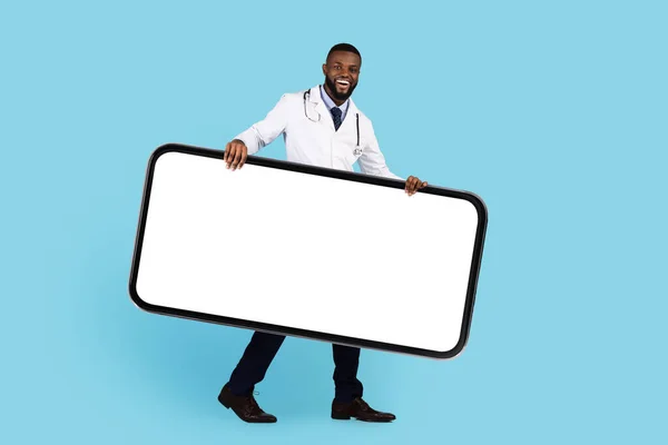 Medical App. Smiling Black Doctor Wearing Uniform Carrying Big Blank Smartphone, Cheerful African American Physician Advertising New Mobile App Or Website While Posing Over Blue Background, Mockup