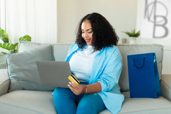 Happy Black Plus Size Woman Shopping Via Laptop And Credit Card Buying New Clothes And Making Online Payment Sitting On Sofa At Home. Internet Banking And Ecommerce Concept