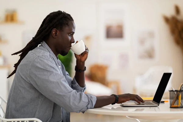 Remote Work. Black Freelancer Guy Working With Laptop And Drinking Coffee While Sitting At Desk In Home Office, African American Man With Dreadlocks Typing On Computer Keyboard, Side View Shot