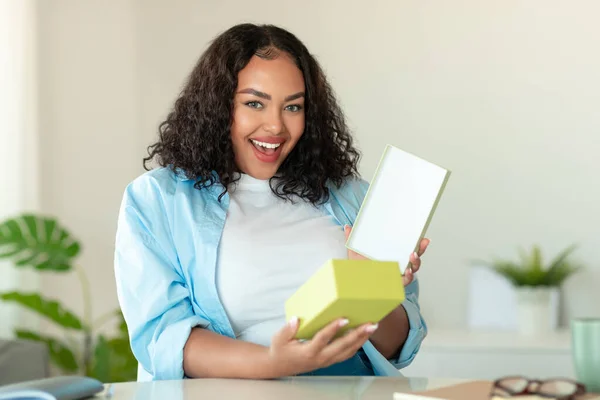 Excited Black Lady Opening Cardboard Box Receiving Parcel After Successful Shopping Sitting At Desk Smiling To Camera At Home. Selective Focus. Presents Delivery Service Concept