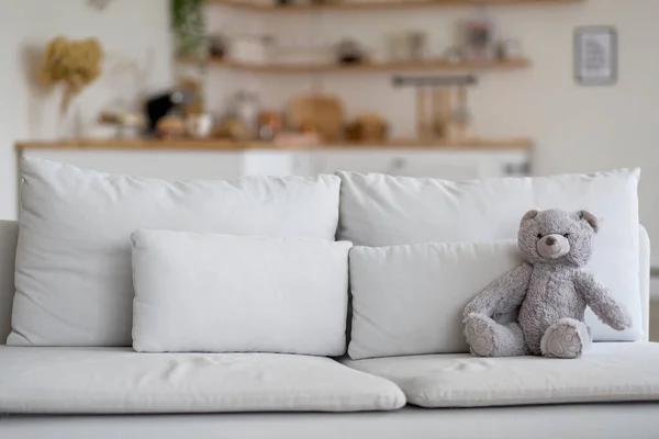 Grey teddy bear sitting on sofa in empty cozy white living room. Lonely teddy bear sitting alone on couch at home, copy space. Lost child, International missing childrens day concept