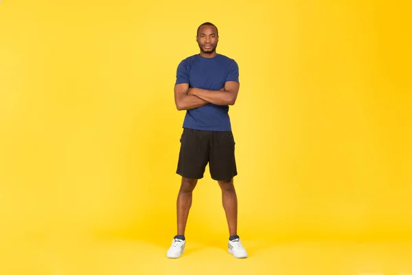 Sport Motivation. Determined African American Sportsman Crossing Hands Posing Looking At Camera Standing On Yellow Studio Background, Wearing Fitwear. Fitness Workout Concept. Full Length