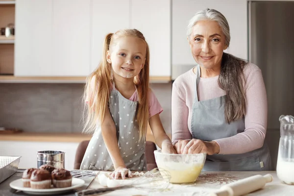 Granddaughter and grandma prepare cookies. Glad european little girl and elderly female in aprons make dough with flour, have fun in kitchen interior. Chefs cooking together at home, household chores