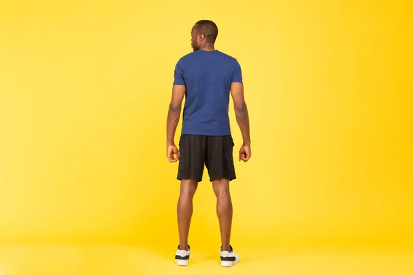 Rear View Of Unrecognizable Sporty Black Man Posing Standing Back To Camera Wearing Fitwear Over Yellow Studio Background. Fitness Workout Concept. Full Length Shot