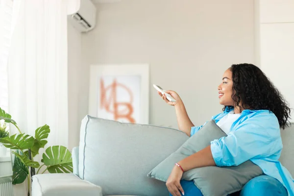 Happy Black Female Turning On AC With Remote Controller Sitting On Couch At Home. Air Conditioning And Climate Control Technologies Concept. Selective Focus