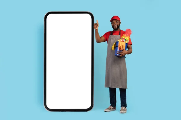 Cleaning Company Ad. Smiling Black Male Cleaner Standing Near Big Blank Smartphone With White Screen, Holding Basket Of Detergents And Showing Thumb Up While Posing Over Blue Background, Mockup
