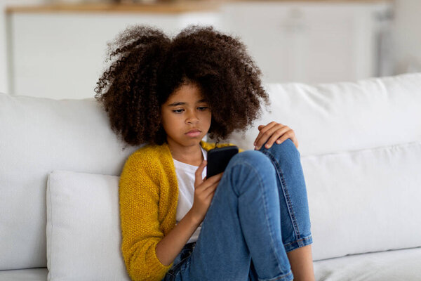 Upset pretty black teen girl with nice bushy hair sitting on sofa at home, using smartphone, kid checking social media, feeling lonely or bored, miss her friends, copy space. Loneliness in children