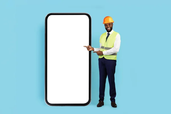 Construction Offer. Smiling Black Male Engineer Pointing At Huge Blank Smartphone With White Screen, African American Civil Engineer In Safety Vest And Hard Hat Recommending New Mobile App, Mockup