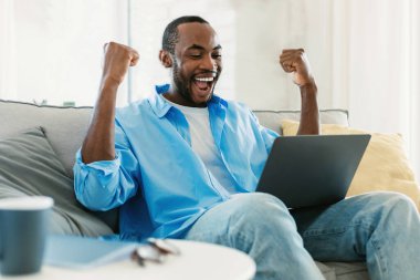 Portrait of overjoyed black man gesturing YES in front of laptop, celebrating achievement, sitting on couch at home, copy space. Remote job, online win concept