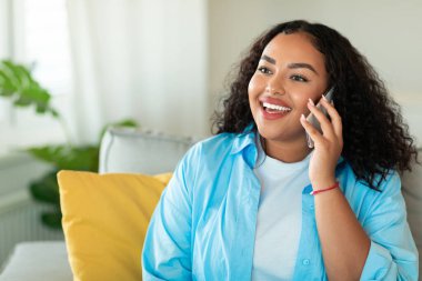 Cheerful African American Lady Talking On Phone Chatting Sitting On Couch At Home. Modern Mobile Communication, Technology And Gadgets Lifestyle Concept