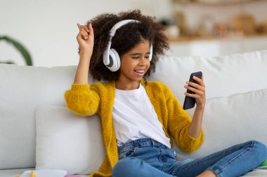 joyful african american school girl with bushy hair sitting on couch at home, using wireless stereo headphones and looking at cell phone screen, raising hand up, listening to music online, copy space