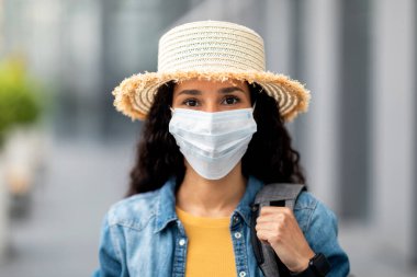 Closeup of long-haired brunette millennial woman in protective face mask carrying backpack, wearing summer straw hat, going vacation while coronavirus pandemic, copy space