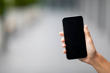 Unrecognizable female hand holding modern cell phone with black empty screen, mockup, selective focus on smartphone, blurred background, panorama with copy space, mobile app, online offer, concept