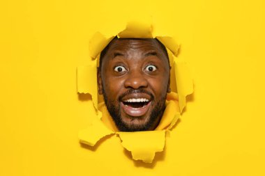 Wow. Overjoyed black man with open mouth breaking and looking through hole in slit yellow paper, copy space. Funky guy amazed of big sale or great news