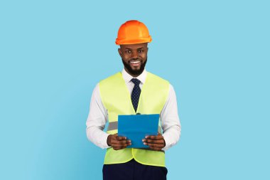 Professions Concept. Handsome Black Engineer In Hard Hat And Vest Holding Clipboard And Smiling At Camera, Portrait Of Young African American Construction Manager Posing On Blue Studio Background