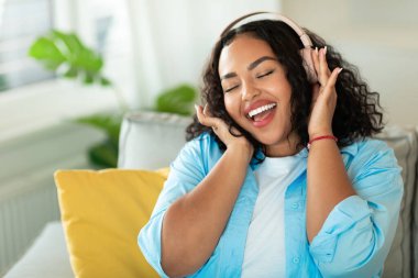 Joyful African American Female Wearing Wireless Earphones Listening To Music Enjoying Song Sitting On Sofa At Home, Posing With Eyes Closed. Great Playlist And Musical Application Concept