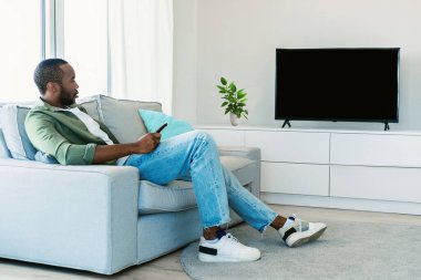 Rest and relax. Black man sitting on the sofa in living room, watching television show, looking at blank plasma tv on the wall with isolated empty monitor, mock up