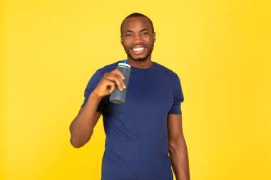 Fitness And Hydration. Cheerful Sporty Black Man Drinking Water Holding Bottle Posing Standing Over Yellow Studio Background. Sport And Healthy Lifestyle Concept