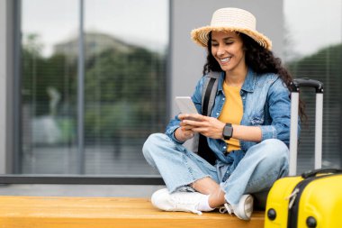 Closeup of cheerful young long-haired brunette lady in straw hat with backpack and suitcase tourist using smartphone, sitting on bench outdoors, booking hotel on Internet, copy space