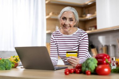 Online Shopping. Happy Elderly Lady Using Laptop And Credit Card In Kitchen While Cooking Food At Home, Smiling Senior Woman Ordering Groceries From Internet Or Making Payments, Copy Space