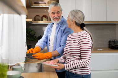 Helping Hand. Happy Senior Man Helping Wife With Dishes In Kitchen, Smiling Elderly Woman And Her Husband Washing And Wiping Plates Together, Making Domestic Chores At Home, Free Space