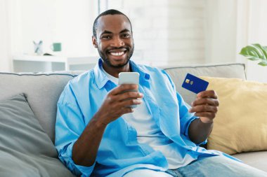 Happy african american man shopping online using smartphone application and credit card, sitting on sofa at home. E-commerce app and internet banking concept