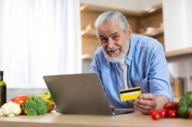 Online Shopping. Smiling Elderly Man Using Laptop And Credit Card In Kitchen While Cooking Lunch At Home, Happy Senior Male Ordering Groceries From Internet Or Making Payments, Copy Space