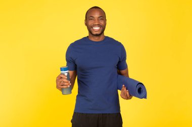 Fitness Workout. Athletic Black Male Holding Water Bottle And Gymnastics Mat Posing Over Yellow Background In Studio, Smiling To Camera. Sport Motivation And Healthy Hydration Concept