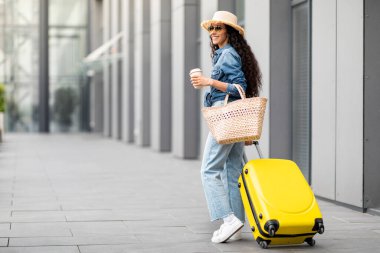 Cheerful young woman in casual outfit summer straw hat and sunglasses walking by modern airport with yellow luggage, holding paper cup, drinking coffee, panorama with copy space, back view