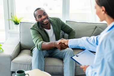 Gratittude for work. Happy black man handshaking with his psychologist after therapy session meeting, sitting on couch at office and smiling
