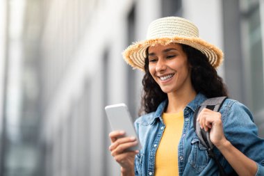 Closeup of cheerful young long-haired brunette lady in straw hat with backpack tourist using smartphone outdoors, looking at cell phone screen and smiling, booking hotel on Internet, copy space