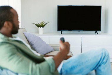 Relaxed black guy watching TV holding remote control and switching channels on flatscreen plasma television set with blank screen, sitting on sofa at home, focus on tv