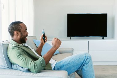 Excited black man watching sport television channel, cheering favorite team, sitting in front of blank plasma screen at home. Mockup, space for your design