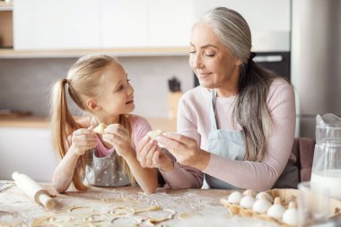 Smiling old grandmother in apron teaching little granddaughter making dough, mold cookies, prepare food in minimalist kitchen interior. Household chores together, cooking lesson at home, free space