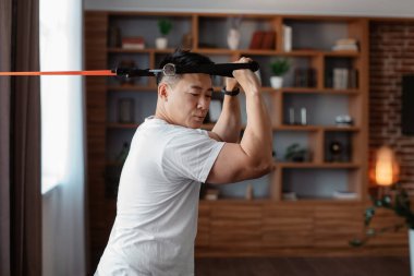 Domestic workout concept. Sporty asian mature man exercising with resistance rubber band, training in living room at home, free space. Workout equipment and sporty lifestyle concept