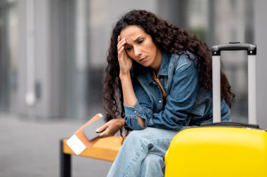 Canceled or delayed flight, difficulties while travelling. Upset young woman tourist with big suitcase sitting on bench outside airport, holding documents, phone, waiting for her flight, copy space